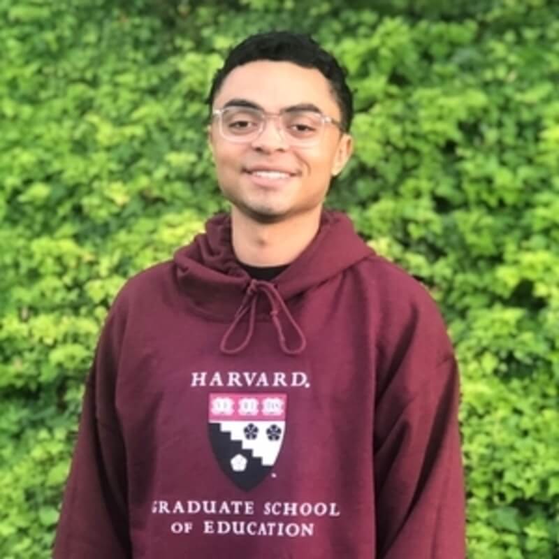 Austin Martin wearing a crimson Harvard sweatshirt standing in front of green tree leaves