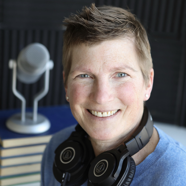 A headshot of alumna Amy Hermon - she has close cropped hair and is facing the camera with a wide smile. Her headphones are casually worn around her neck and a microphone is pictured in the background.