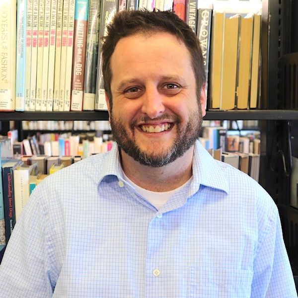 A headshot of School of Information Sciences Calvin Battles. He is pictured in front of library shelves lined with books. He has short hair and facial hair and is smiling at the camera.
