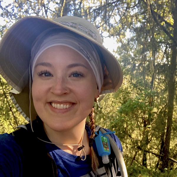 SIS student Emily is pictured in front of trees during a hike. She is wearing a wide-brimmed hat and a wide smile. 