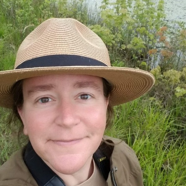 SIS Alumna Jodi Coalter is pictured outdoors in front of a wooded area with a pond in the foreground. She is wearing a wide-brimmed hat.