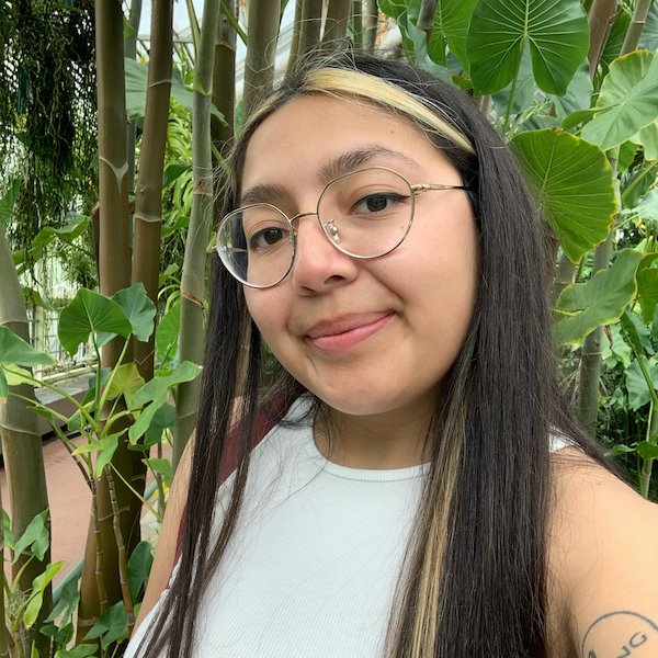SIS student Selena Aguilera is pictured wearing a white top. She has glasses and long hair with light streaks in the front. She is pictured in front of vibrant green plants.