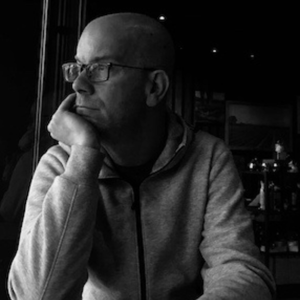 In a black and white image, Browning looks pensively out of a window. He has a shaved head and is wearing glasses and a casual hooded shirt.