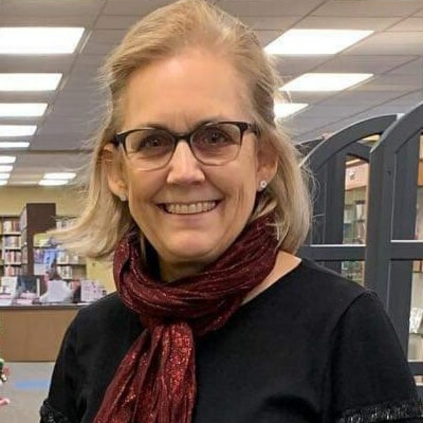 A headshot of School of Information Sciences alumna Sue Todd. She smiling at the camera and is wearing a red scarf and glasses and has short blonde hair that is pulled back at the temples. 