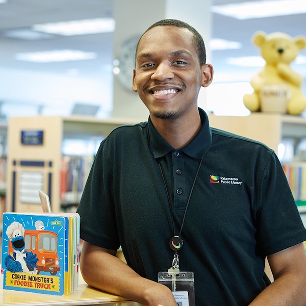 Theo Gill, a young Black man with short hair and a wide smile is pictured in the library. He is wearing a black polo shirt. 