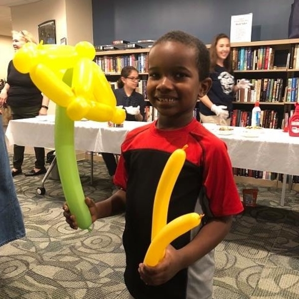 little boy smiling in front of a row of library books holding yellow and green balloons